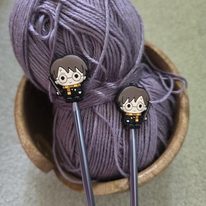May include: Two knitting needles with decorative toppers featuring a cartoon character with brown hair, round glasses, a lightning bolt scar, and a black and yellow scarf. The needles are inserted into a ball of purple yarn inside a wooden bowl.