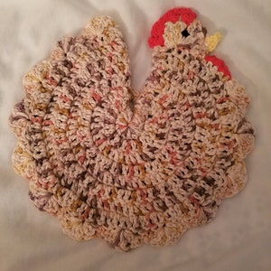 May include: A crocheted chicken dishcloth in shades of brown, pink, and cream. The chicken has a red comb and a yellow beak.