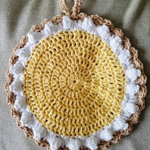 May include: A crocheted pot holder in the shape of a sunflower with a yellow center, white petals, and a brown outer edge. The pot holder has a loop for hanging.