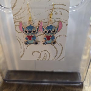 Puede incluir: Pendientes colgantes con un personaje de dibujos animados azul sosteniendo un coraz&oacute;n rojo. Los pendientes tienen ganchos dorados y orejas rosas. Se exhiben en una tarjeta blanca con un patr&oacute;n dorado.