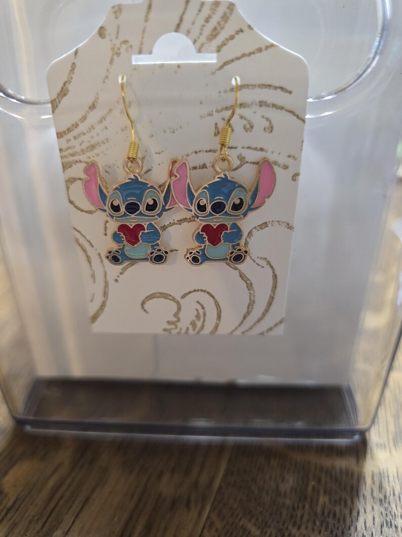 Puede incluir: Pendientes colgantes con un personaje de dibujos animados azul sosteniendo un coraz&oacute;n rojo. Los pendientes tienen ganchos dorados y orejas rosas. Se exhiben en una tarjeta blanca con un patr&oacute;n dorado.