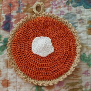 May include: A crocheted orange and beige pot holder shaped like a pie with a white crocheted center.