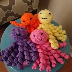 May include: Four crocheted octopus toys in bright colors including purple, pink, orange, and yellow. Each octopus features a round head with a stitched smiling face and eight spiraled, curly tentacles extending outward from the base.