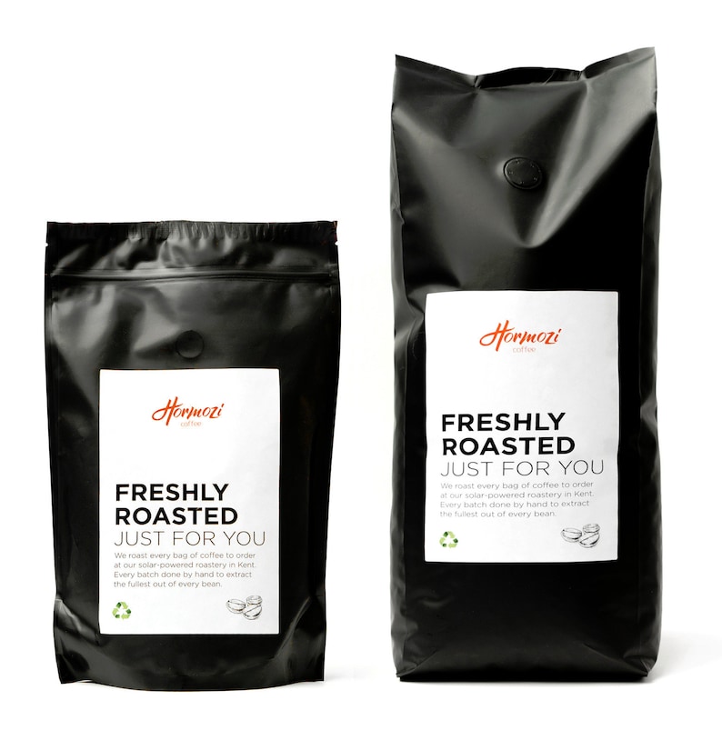 K&ouml;nnte beinhalten: Zwei schwarze Kaffeet&uuml;ten mit wei&szlig;en Etiketten. Auf den Etiketten steht "Hormozi Coffee" und "FRESHLY ROASTED JUST FOR YOU". Die T&uuml;ten haben eine matte Oberfl&auml;che und ein kleines, kreisf&ouml;rmiges Ventil. Der Text erw&auml;hnt auch das R&ouml;sten mit Solarenergie.