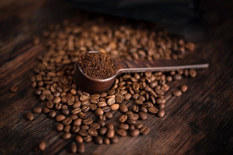 K&ouml;nnte beinhalten: Ein h&ouml;lzerner L&ouml;ffel, gef&uuml;llt mit gemahlenem Kaffee, liegt auf einem Bett aus ger&ouml;steten Kaffeebohnen. Der L&ouml;ffel und die Bohnen befinden sich auf einer dunklen Holzoberfl&auml;che. Das Bild hebt die satten Braunt&ouml;ne von Kaffee und Holz hervor.