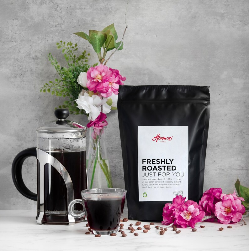K&ouml;nnte beinhalten: Ein schwarzer Beutel mit frisch ger&ouml;stetem Kaffee mit einem wei&szlig;en Etikett mit der Aufschrift "FRESHLY ROASTED JUST FOR YOU". Eine French Press, eine transparente Glas-Tasse und eine Vase mit rosa und wei&szlig;en Blumen sind ebenfalls im Bild. Kaffeebohnen sind verstreut.