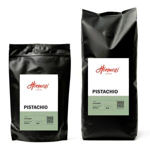 May include: Two black coffee bags with white labels. The labels read "Hormozi coffee" in red script and "PISTACHIO" in bold black letters. The bottom of the label has the words "FLAVOURED", "ROAST COLOUR", and "STRENGTH".