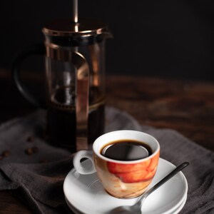K&ouml;nnte beinhalten: Eine Kaffeetasse auf einer wei&szlig;en Untertasse mit einem L&ouml;ffel, auf einer grauen Serviette. Im Hintergrund befindet sich eine French-Press-Kaffeemaschine. Die Tasse hat ein orange-wei&szlig;es Design und ist mit dunklem Kaffee gef&uuml;llt.