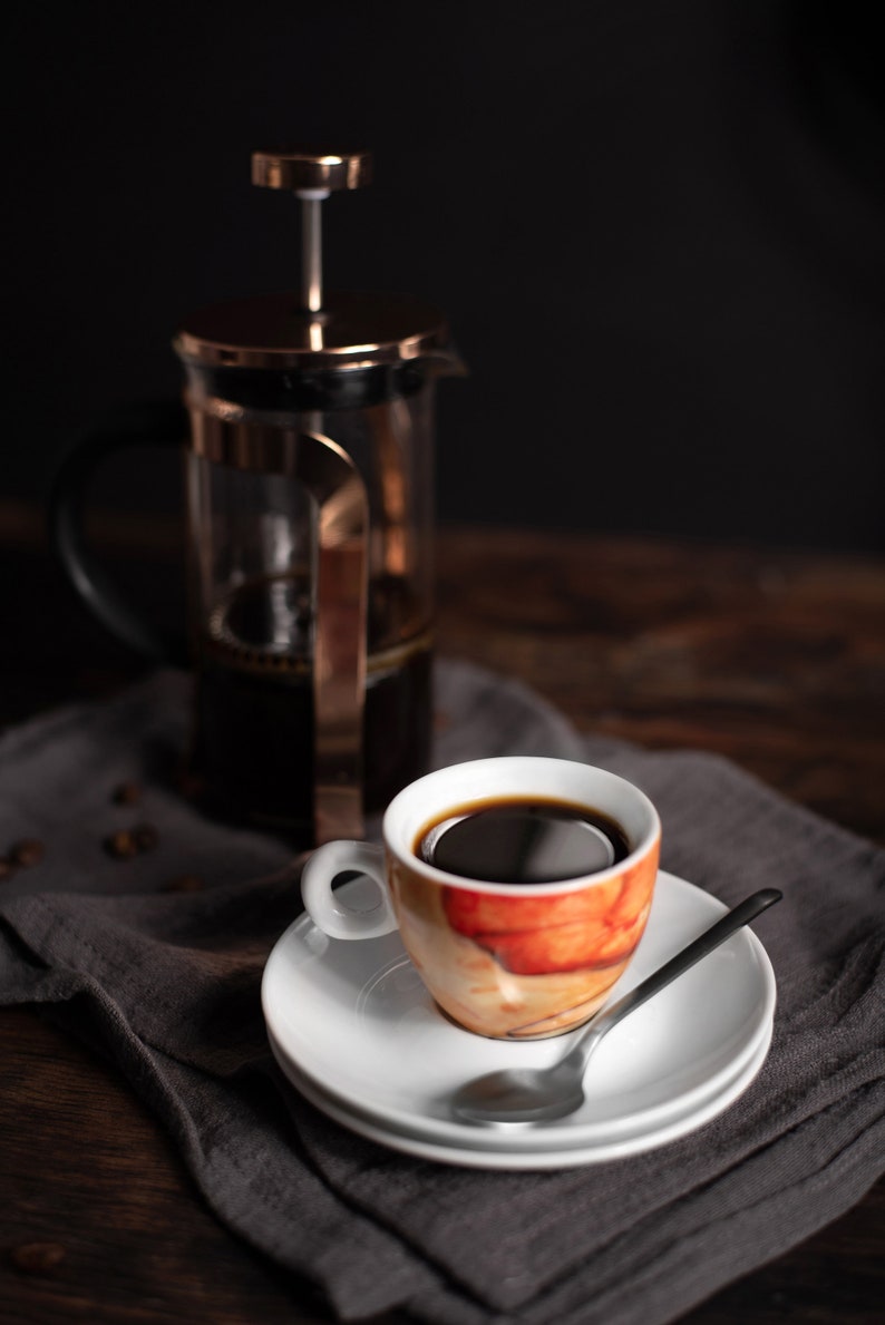 K&ouml;nnte beinhalten: Eine Kaffeetasse auf einer wei&szlig;en Untertasse mit einem L&ouml;ffel, auf einer grauen Serviette. Im Hintergrund befindet sich eine French-Press-Kaffeemaschine. Die Tasse hat ein orange-wei&szlig;es Design und ist mit dunklem Kaffee gef&uuml;llt.