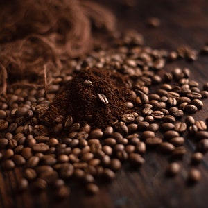 K&ouml;nnte beinhalten: Nahaufnahme von Kaffeebohnen und gemahlenem Kaffee auf einer dunklen Holzoberfl&auml;che. Die ger&ouml;steten Kaffeebohnen umgeben einen kleinen Haufen gemahlenen Kaffees, mit ein paar Bohnen obenauf. Das Bild hat eine warme, einladende Ausstrahlung.