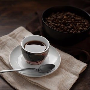 K&ouml;nnte beinhalten: Eine Tasse Kaffee auf einer wei&szlig;en Untertasse mit einem L&ouml;ffel, auf einer beigen Leinen-Serviette. Eine schwarze Sch&uuml;ssel mit Kaffeebohnen befindet sich im Hintergrund. Die Szene ist auf einer dunklen Holzoberfl&auml;che.