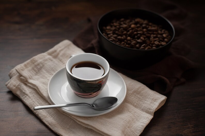 K&ouml;nnte beinhalten: Eine Tasse Kaffee auf einer wei&szlig;en Untertasse mit einem L&ouml;ffel, auf einer beigen Leinen-Serviette. Eine schwarze Sch&uuml;ssel mit Kaffeebohnen befindet sich im Hintergrund. Die Szene ist auf einer dunklen Holzoberfl&auml;che.