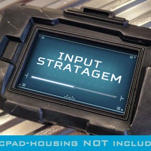 May include: A black rectangular electronic device with a screen displaying the text INPUT STRATAGEM in white capital letters. The device features a rugged, dark frame with visible scuff marks and a progress bar at the bottom of the screen.