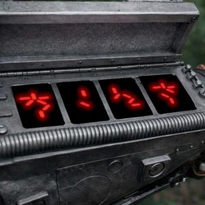 Könnte beinhalten: Ein verwittertes, metallisch-graues Requisit aus dem Film Predator. Es hat einen Klappdeckel und vier rechteckige Displays. Jedes Display zeigt leuchtend rote Symbole auf schwarzem Hintergrund. Das Requisit hat ein strukturiertes, industrielles Design.
