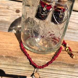 May include: A set of handmade jewelry featuring small glass bottles filled with black crystals and topped with cork stoppers. The bottles are suspended from gold-colored wire wrapped in red thread and shaped into hearts. The necklace features a similar bottle pendant.