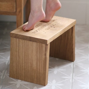 May include: A wooden step stool with the words "Erin's Toothbrushing School" engraved on the top. A child's feet are on the top of the stool.