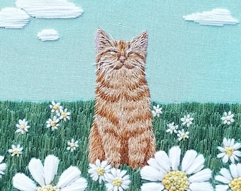 May include: Embroidered hoop art featuring a ginger cat sitting in a field of white daisies. The cat is smiling and has a light blue background.