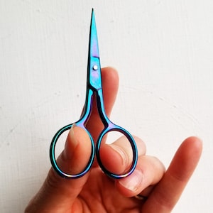 May include: A pair of small, iridescent blue and green scissors with a silver handle. The scissors are held in a hand with the blades open.
