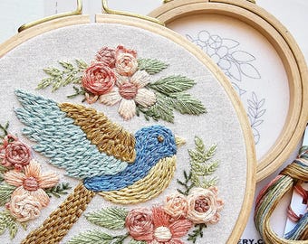 May include: A bluebird embroidered on a white fabric with pink and white flowers. The embroidery is in a wooden hoop. The image also shows a card with floss colors and numbers.