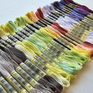 May include: A collection of 20 skeins of embroidery floss in various shades of grey, brown, yellow, green, and peach. The floss is arranged in rows, with the darkest shades at the bottom and the lightest shades at the top. Each skein is labeled with a number from 01 to 20.
