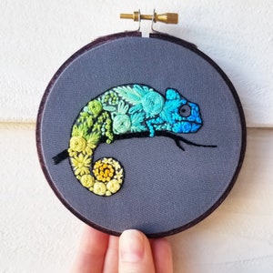 May include: A colourful embroidered chameleon on a grey fabric background. The chameleon is made of green, yellow and blue embroidery floss. The chameleon is curled up in a circle.