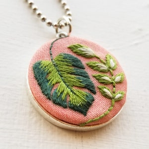 Puede incluir: Un collar con un colgante de tela rosa y una cadena plateada. El colgante tiene una hoja verde bordada y un tallo blanco con peque&ntilde;as hojas.