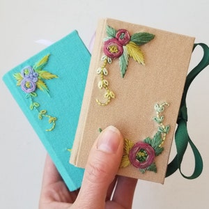 May include: Two small, embroidered books. One is teal, the other tan. Both feature floral embroidery in shades of purple, pink, green, and yellow. A green ribbon is attached to the tan book. The books are held in a hand.