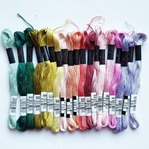 Pode incluir: Uma cole&ccedil;&atilde;o de 18 novelos de fio para bordar em v&aacute;rios tons de verde, amarelo, rosa, vermelho e roxo. O fio est&aacute; disposto em uma fila, com as etiquetas voltadas para o observador. As etiquetas s&atilde;o pretas com texto branco e incluem o nome da marca "cosmo" e um n&uacute;mero.