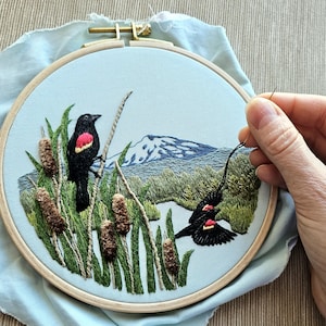 May include: Embroidered artwork featuring two black birds with red wing markings, perched on reeds and cattails. A mountain range and landscape are in the background. The embroidery is set in a wooden hoop on a light blue fabric.
