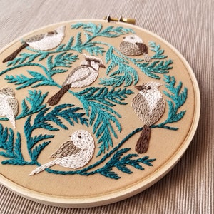 以下が含まれることがあります： ベージュの背景にターコイズグリーンの葉と白い鳥が刺繍されたフープアート。鳥はフープの周りに円形に配置されています。