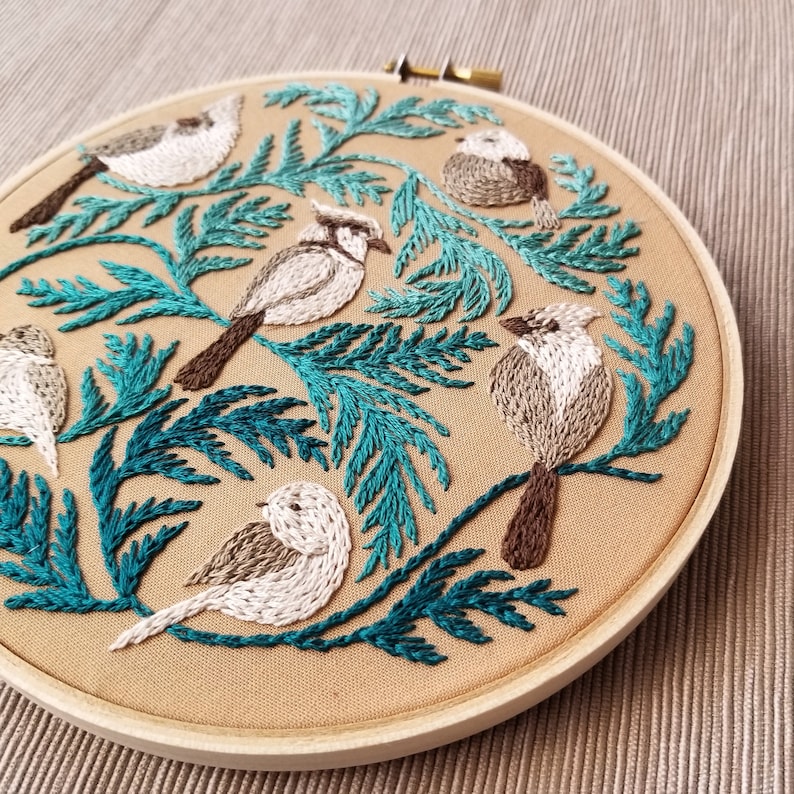 以下が含まれることがあります： ベージュの背景にターコイズグリーンの葉と白い鳥が刺繍されたフープアート。鳥はフープの周りに円形に配置されています。