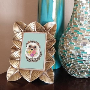 May include: A gold-coloured picture frame with a light blue background and a picture of a brown and white hedgehog holding a bouquet of pink, purple and yellow flowers.