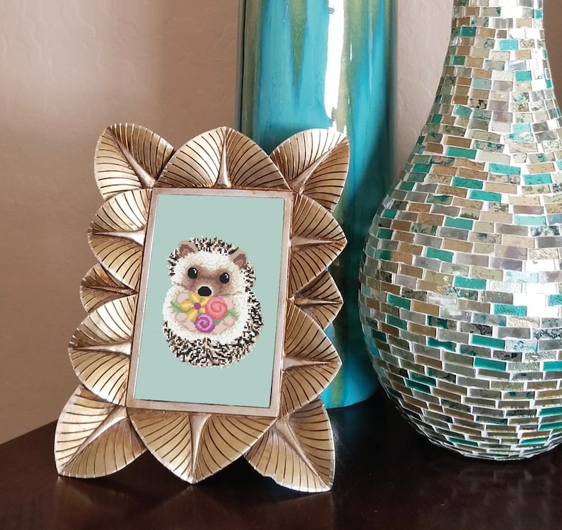 May include: A gold-coloured picture frame with a light blue background and a picture of a brown and white hedgehog holding a bouquet of pink, purple and yellow flowers.