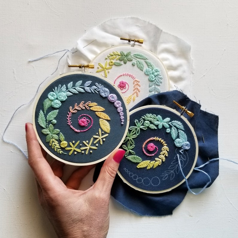 K&ouml;nnte beinhalten: Drei Stickrahmen mit bunten Blumenmustern. Die Rahmen liegen auf einem dunkelblauen Stoffhintergrund. Die Muster sind alle unterschiedlich, aber sie alle haben ein Spiralmuster.