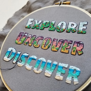 May include: Embroidered hoop art featuring the words "Explore, Uncover, Discover" in colourful thread. Each word is decorated with a different landscape scene.