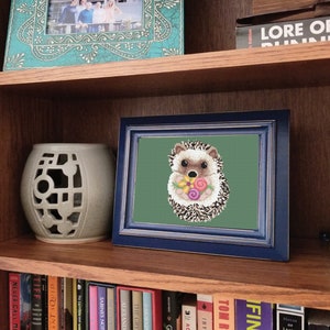 May include: A framed cross-stitch embroidery of a hedgehog holding a bouquet of flowers. The hedgehog is brown and white with a pink nose. The flowers are pink, yellow, and orange. The background is a dark green colour.