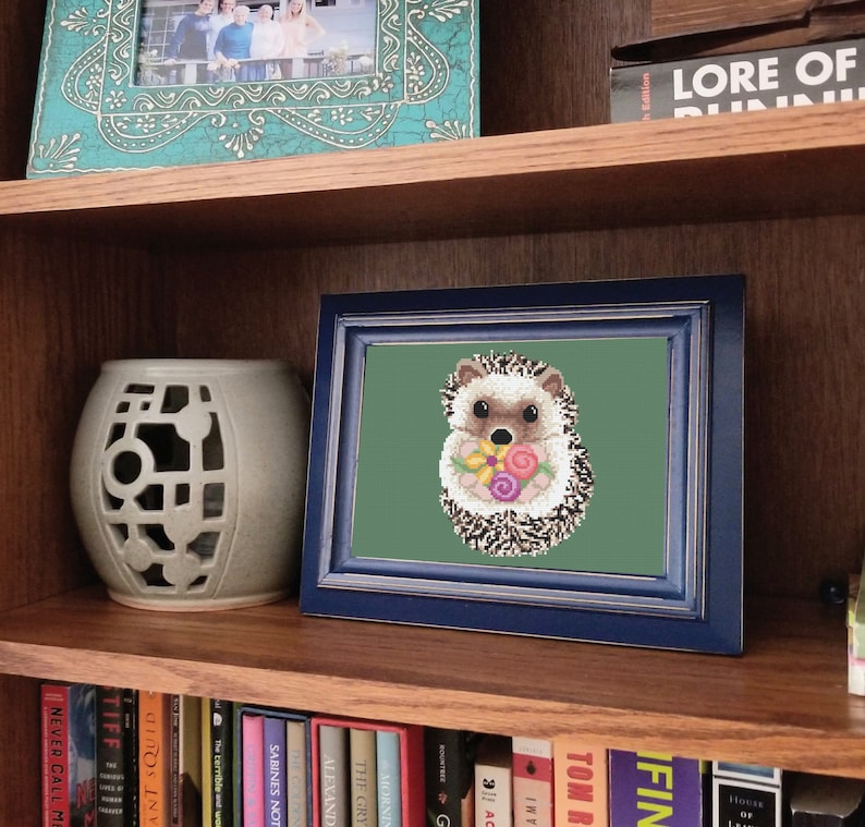 May include: A framed cross-stitch embroidery of a hedgehog holding a bouquet of flowers. The hedgehog is brown and white with a pink nose. The flowers are pink, yellow, and orange. The background is a dark green colour.