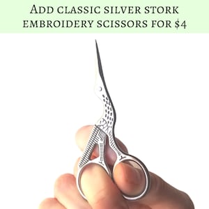 Puede incluir: Tijeras de bordado de cig&uuml;e&ntilde;a plateada con un dise&ntilde;o detallado. Las tijeras est&aacute;n abiertas y se sostienen en una mano.