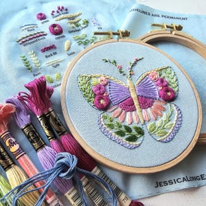 May include: A completed embroidery project featuring a butterfly design in a wooden hoop. The butterfly is stitched with purple, pink, and green thread on a light blue fabric. Embroidery floss in various colors and a fabric with stitch examples are also visible.