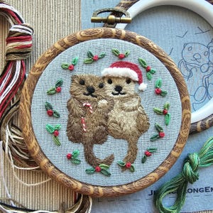 May include: Embroidered hoop art featuring two otters, one wearing a Santa hat, surrounded by holly. The otters are brown and tan, with a candy cane. The hoop is wood-toned, and the background is light blue.