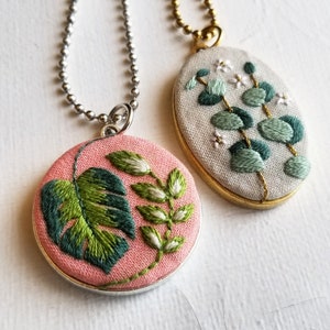 Puede incluir: Dos collares bordados con dise&ntilde;os florales. Un collar tiene un fondo rosa con una hoja de monstera verde y flores blancas. El otro collar tiene un fondo blanco con hojas de eucalipto verdes y flores blancas.