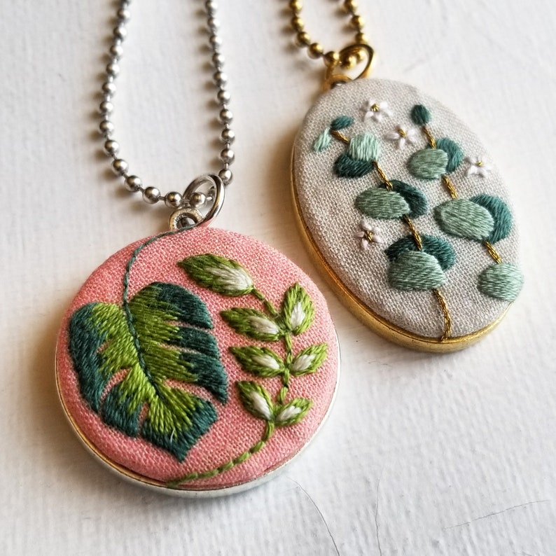 Puede incluir: Dos collares bordados con dise&ntilde;os florales. Un collar tiene un fondo rosa con una hoja de monstera verde y flores blancas. El otro collar tiene un fondo blanco con hojas de eucalipto verdes y flores blancas.