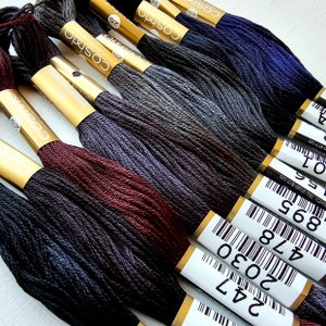 May include: Close-up of several skeins of Cosmo embroidery floss in various shades of gray, burgundy, and navy blue. Each skein is labeled with a white sticker and gold foil accents. The floss is tightly wound and ready for use.