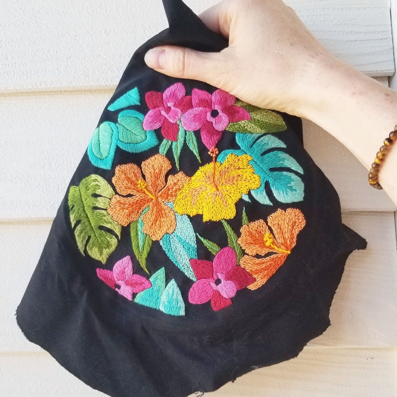 May include: Black fabric with embroidered tropical flowers in shades of pink, yellow, green, and blue. The flowers are detailed and realistic.