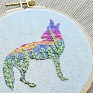 May include: An embroidery hoop featuring a colorful coyote design. The coyote is filled with a sunset gradient of pink, yellow, and blue. Green cacti are embroidered around the coyote's body. The hoop is made of wood and the fabric is a light beige.