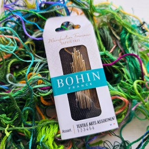 May include: A package of Bohin Textile Arts Assortment needles, size 1-2-3-4-5-6, in a white box with black and blue accents. The box is surrounded by colorful embroidery floss.