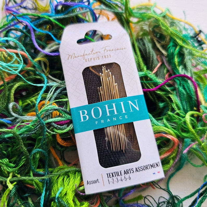 May include: A package of Bohin Textile Arts Assortment needles, size 1-2-3-4-5-6, in a white box with black and blue accents. The box is surrounded by colorful embroidery floss.