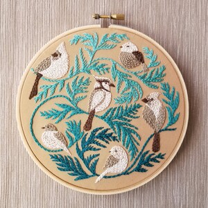 以下が含まれることがあります： 5羽の鳥が葉のついた枝にとまっている刺繍のフープアート。鳥はベージュと白で、茶色のアクセントが付けられています。枝は鮮やかなターコイズブルーです。