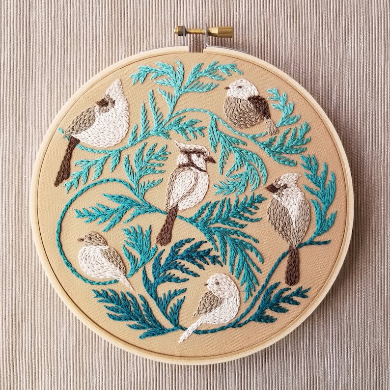 以下が含まれることがあります： 5羽の鳥が葉のついた枝にとまっている刺繍のフープアート。鳥はベージュと白で、茶色のアクセントが付けられています。枝は鮮やかなターコイズブルーです。