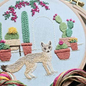 May include: Embroidered artwork featuring a desert fox amidst cacti and flowering plants in terracotta pots. The design includes a light blue background and a decorative arch. The embroidery uses various shades of green, pink, and brown, creating a detailed and textured piece.